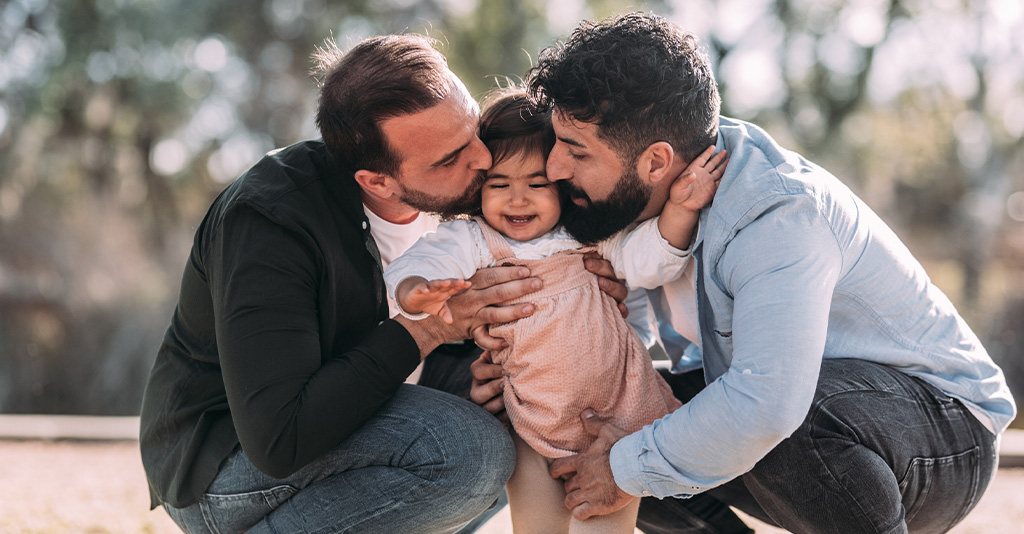 Gay couple kissing their daughter who is smiling amusedly in an urban park.