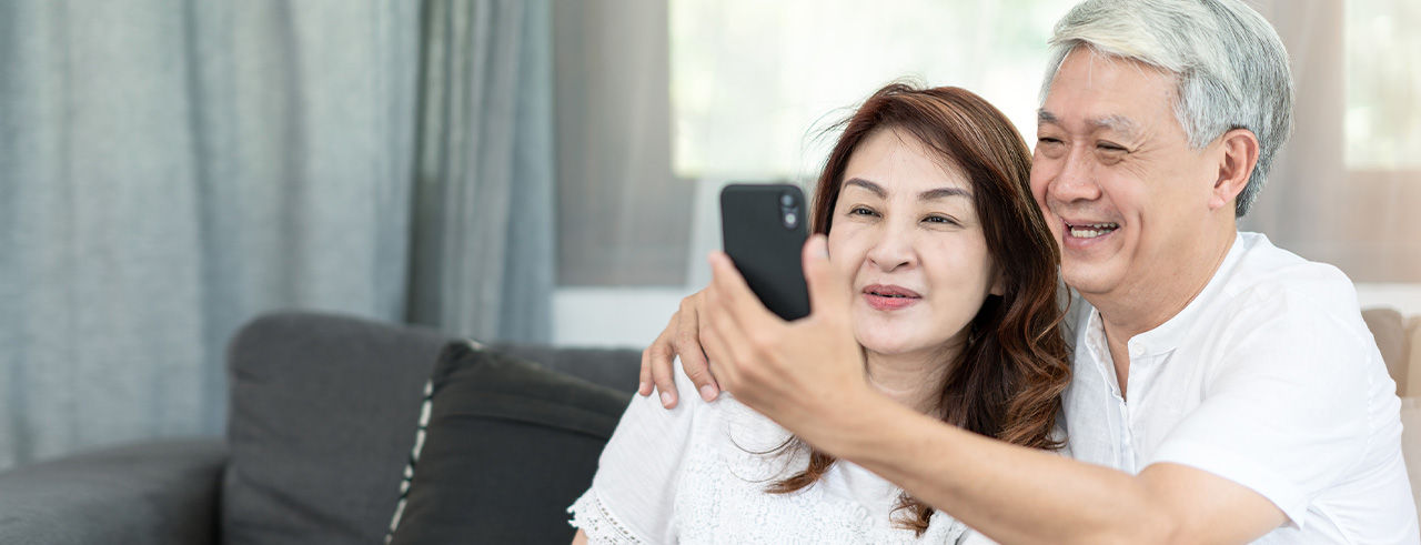 Candid of attractive old asian couple husband and wife use technology sitting on coach at home. Senior parent watching or looking at mobile screen.