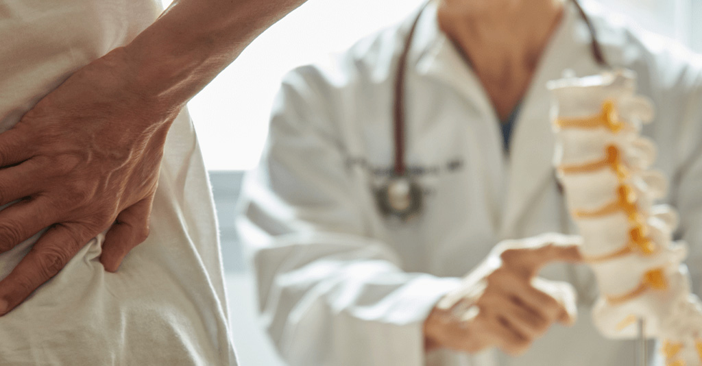 A doctor pointing to a section of the spine while a woman holds her lower back in the foreground.